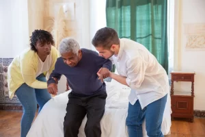 Two caregivers from the best NDIS service provider helping an old man to stand up from the bed.