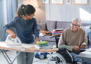 Young caregiver ironing clothes while talking with old guy sitting in the wheel chair.