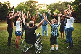 A diverse group of people including a person in wheelchair, holding hands in a circle outdoors, showing a community participation.